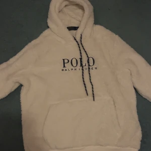 Vit fleecehoodie Polo Ralph Lauren - Supermjuk och fluffig vit hoodie från Polo Ralph Lauren med broderad logga på bröstet och ärmen. Tröjan har huva med snörning och stor känguruficka framtill. Perfekt för kalla dagar när du vill vara både bekväm och snygg.