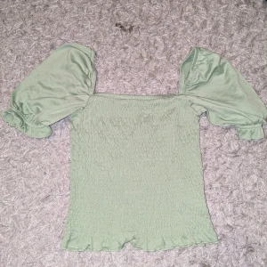 Grön smockad blus med puffärm - Söt ljusgrön blus med smockad kropp och volangkant nertill. Toppen har korta puffärmar med resår och fyrkantig urringning. Perfekt för dig som gillar romantisk och trendig stil.