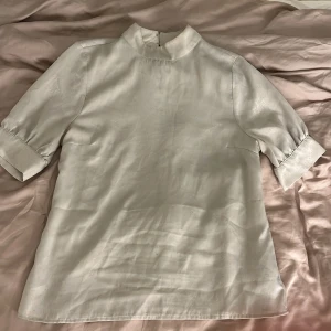 Ljusbeige blus i satin från Gina Tricot - Säljer en ljusbeige blus från Gina Tricot i satinliknande material. Blusen har hög krage, halvlånga ärmar med breda manschetter och dragkedja bak. Perfekt för dig som gillar stilrena och eleganta plagg med en modern touch.
