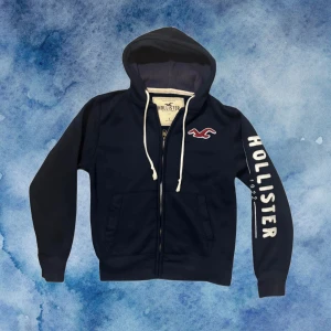 Mörkblå Vintage Hollister Kofta  - Mörkblå hoodie från Hollister med vit dragsko, dragkedja och stor logga på ena ärmen. Broderad röd logga på bröstet och kängurufickor framtill. Koftan är lite nopprig men inget som syns. 