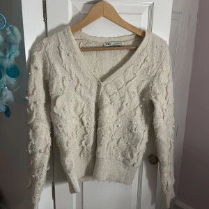 Vit kofta med pärlor från Zara - Superfin vit kofta från Zara med v-ringning och dekorativa pärlor över hela tröjan. Stickad i mjukt material med långärmad passform och ribbade muddar. Perfekt för dig som vill ha en mysig men ändå stylish look. Denna kofta säljer jag för den inte kommer till användning längre, använd den 5 gånger för 1 år sen ‼️Super fin och söt 🥰