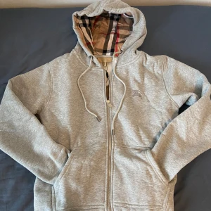 Grå hoodie från Burberry med dragkedja - Snygg grå hoodie från Burberry med klassiskt rutigt foder i huvan och broderad logga på bröstet. Tröjan har dragkedja framtill, snörning i huvan och två fickor. Perfekt för dig som gillar stilrena och exklusiva plagg.