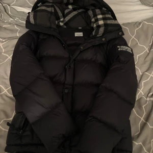 Svart pufferjacka från Burberry - Svart pufferjacka från Burberry med avtagbara ärmar så den kan användas som väst. Jackan har en stor huva med rutigt foder i grått och vitt, samt dragkedja och knappar framtill. Burberry-logga på ärmen och två sidofickor med dragkedja. Priset är ej hugget i sten. Tveka ej om du har några frågor