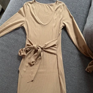 Beige ribbad klänning med knytband - Snygg beige ribbad klänning med lång ärm och v-ringning. Klänningen har ett brett knytband i midjan som ger en fin siluett. Perfekt för dig som gillar stilrena och trendiga plagg.