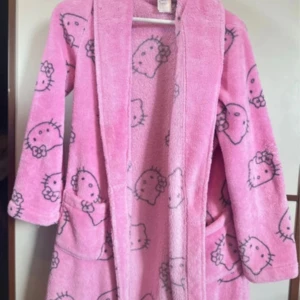 Hello Kitty rosa morgonrock  - Supermysig rosa morgonrock  med Hello Kitty-mönster över hela. Jackan har långa ärmar, stora fickor och en skön krage. Perfekt för dig som älskar gulliga och lekfulla plagg. Sitter löst och är riktigt mjuk mot huden.
