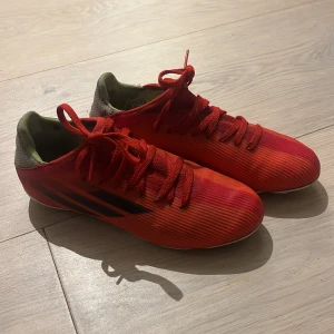 Röda Adidas X fotbollsskor - Säljer ett par röda Adidas X fotbollsskor med svarta detaljer och vita dobbar. Skorna har snörning och en smidig, låg profil med rund tå. Ovandelen är i syntetmaterial och hälen har grå mesh med X-logga. Perfekt för dig som vill sticka ut på planen.