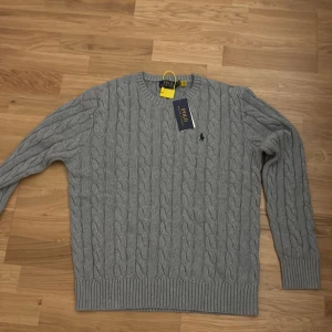 Grå kabelstickad tröja Polo Ralph Lauren - Kabelstickad grå tröja från Polo Ralph Lauren med klassisk rund hals och broderad logga på bröstet. Tröjan har ribbade muddar och är tillverkad i mjuk bomull. Perfekt för lager-på-lager och snygg till jeans eller kjol. Pris ej hugget i sten!