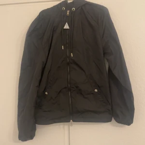 Svart vindjacka från Moncler - Svart vindjacka från Moncler med huva som har röd, vit och blå rand. Jackan har dragkedja framtill, två fickor med tryckknappar och Moncler-logga på ärmen. Materialet är lätt och syntetiskt, perfekt för blåsiga dagar.