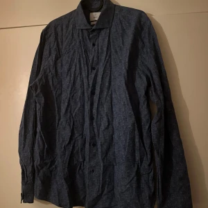 Blå mönstrad skjorta från Bläck - Snygg blå skjorta från Bläck i regular fit med diskret geometriskt mönster. Skjortan har klassisk krage, svarta knappar och långa ärmar. Perfekt för dig som gillar stilrena detaljer och vill ha något som sticker ut lite extra.