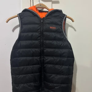 Vändbar dunväst från Hugo BOSS, svart/orange - Vändbar dunväst från Hugo Boss med huva och dragkedja. Ena sidan är svart med orange BOSS-logga, andra sidan är helorange med svart logga. Quiltad design, två fickor på orangea sidan och sportiga ränder vid ärmöppningarna. Perfekt för lager-på-lager och streetstyle. Västen är nästan aldrig använd och är köpt ur en begränsad upplaga som inte säljs längre och som är otroligt sällsynt idag! Kan passa barn mellan 8-11 års ålder beroende på hur stor man är. Storlek 138.