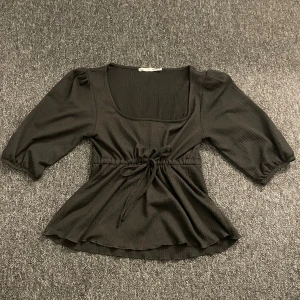 Svart blus med knyt från 157 - Svart blus från 157 med trekvartsärmar, fyrkantig ringning och knyt i midjan. Toppen har lätt rynkade axlar och en flowy passform nedtill. Materialet är ribbat och mjukt, perfekt för en chill och snygg look.