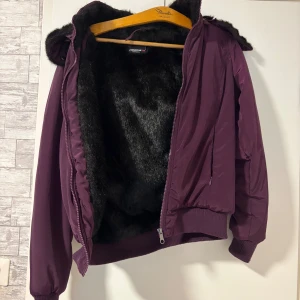 Mörklila pufferjacka från Fishbone - Säljer en mörklila pufferjacka från Fishbone med fluffigt svart foder och huva. Jackan har ribbade muddar, dragkedja framtill och två sidofickor. Perfekt för kalla dagar när du vill vara både varm och snygg. Storlek L.
