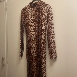 Lång leopardmönstrad klänning - Långärmad klänning med leopardmönster i bruna och beige toner. Klänningen har en figurnära passform, hög hals och är hel-lång. Materialet känns stretchigt och mjukt, perfekt för dig som vill sticka ut med ett djurmönster.