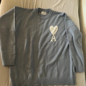 Ljusblå stickad tröja från Ami - Snygg ljusblå stickad tröja från Ami Alexandre Mattiussi med vit hjärtdetalj och bokstaven A på bröstet. Tröjan har rund hals, ribbade muddar och är långärmad. Perfekt för dig som gillar stilrena och trendiga plagg med en twist.