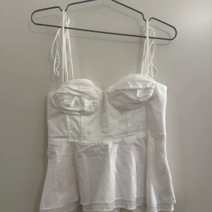 Vit korsett-topp med snörning - Supersnygg vit korsett-topp från Zara med tunna axelband som kan knytas. Toppen har formade kupor och en volang nedtill som ger en feminin vibe. Smockad rygg för extra komfort och passform. Perfekt för dig som vill ha en trendig och chic look. Skriv vid frågor!☺️