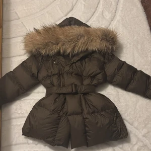 Brun dunjacka med bälte - Snygg brun dunjacka med stor huva och fluffig päls. Jackan har bälte i midjan för en schysst siluett och långa ärmar. Perfekt för höst.