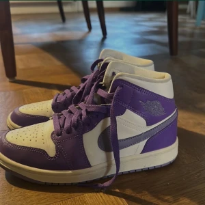 Nike Air Jordan 1 lila/vit EUR 37.5 - Nike Air Jordan 1 sneakers i lila och vitt med klassisk high-top siluett. Skorna har lila snörning, perforerad tå och ikonisk Air Jordan-logga på sidan. Swoosh i lila och vit, samt platt sula i lila. Tillverkade i skinn och syntetmaterial. 