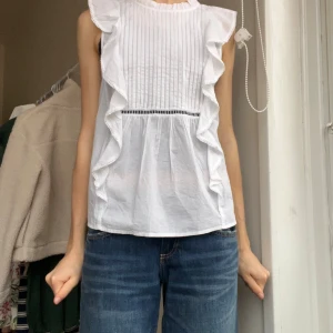 Vit volangblus från BikBok -  vit blus från BikBok med volanger längs sidorna och plisserad framsida. Ärmlös modell med liten ståkrage och dekorativ spetskant. Luftig och lätt i bomull. 