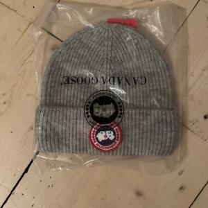 Grå  mössa från Canada Goose - Snygg grå ribbstickad mössa från Canada Goose med klassisk uppvikt kant och ikonisk logopatch framtill. Perfekt för kalla dagar och ger en clean look. Materialet är mjukt och värmande ull.