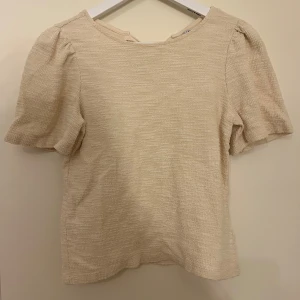 Ljus beige topp med knyt från Holly & Whyte - Superfin ljus beige topp från Holly & Whyte med korta puffärmar och snygg struktur i tyget. Baksidan har en söt knytning vid nacken och en liten öppning. Perfekt till jeans eller kjol. Aldrig använt den så som ny.