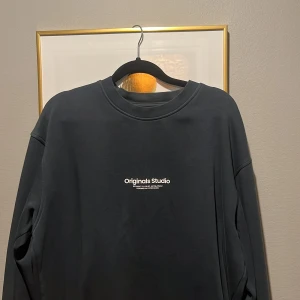 Svart sweatshirt från Originals Studio - Mörk, svart sweatshirt från Originals Studio med vit text på bröstet. Tröjan har rund halsringning och långa ärmar. Enkel och clean design som passar perfekt till jeans eller joggers.