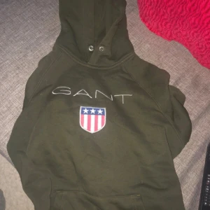 Mörkgrön hoodie från GANT - Snygg mörkgrön hoodie från GANT med broderad logga och emblem på bröstet. Tröjan har huva med snörning och klassisk känguruficka framtill. Perfekt för dig som gillar stilren streetwear med sportig touch.