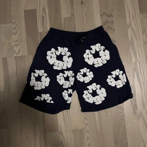 Mörkblå shorts med blommigt tryck - Snygga mörkblå shorts med stora vita blommor tryckta över hela tyget. Modellen har elastisk midja med snörning för skön passform. Perfekta för sommarens chill dagar och har en loose fit som ger en avslappnad vibe.