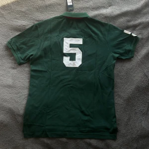 Grön Polo Ralph Lauren Milan piké - Snygg grön pikétröja från Polo Ralph Lauren med stora gula broderier, MILAN-text och emblem på bröstet. Klassisk krage, korta ärmar och vita siffror på ryggen och ena ärmen. Materialet är mjuk bomull och passformen är normal.