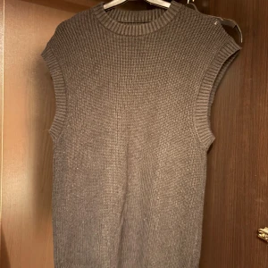 Svart stickad väst med ribbade kanter - En Svart stickad väst med rund hals och ribbade kanter vid ärmhål och halsringning. Västen har en rak och enkel passform, perfekt att styla över en skjorta eller t-shirt för en trendig lager-på-lager-look.