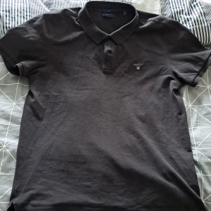 Svart pikétröja från GANT XL - Svart pikétröja från GANT i klassisk stil med krage och korta ärmar. Tröjan har GANT-logga broderad på bröstet och är tillverkad i mjuk bomull. Passformen är normal och modellen är regular fit. Perfekt för en clean och enkel look.