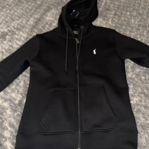 Svart hoodie från Polo Ralph Lauren - Svart hoodie från Polo Ralph Lauren med dragkedja, huva och snörning. Klassisk logga broderad på bröstet och två stora fickor framtill. Skön passform och perfekt för en chill look. Materialet är mjukt och känns riktigt nice knappt använt och bra pris. Pris kan diskuteras vid snabb affär 