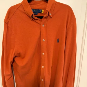 Ralph Lauren skjorta  - Snygg orange skjorta från Polo Ralph Lauren i featherweight mesh. Klassisk krage, långa ärmar och knappar framtill. Broderad blå logga på bröstet ger en stilren touch. Perfekt för dig som gillar färg och vill sticka ut lite extra.