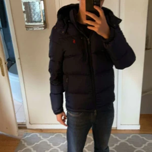 Mörkblå pufferjacka från Polo Ralph Lauren - Säljer en mörkblå pufferjacka från Polo Ralph Lauren med huva och dragkedja. Jackan har två sidofickor med dragkedja och ett rött Polo-logo på bröstet. Materialet är glansigt och syntetiskt, perfekt för kalla dagar. Passformen är normal och jackan är långärmad.
