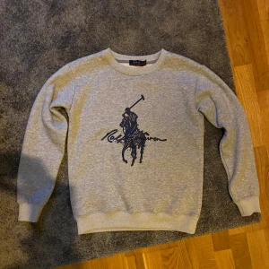 Grå sweatshirt från Polo Ralph Lauren - Säljer en grå sweatshirt från Polo Ralph Lauren med stort mörkblått logotryck på bröstet. Tröjan har rund halsringning, ribbade muddar och är tillverkad i mjuk bomullsmix. Perfekt för en chill och stilren look.