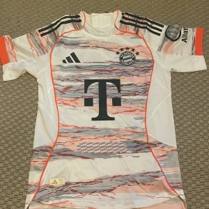 Bayern München Musiala Adidas tröja S - FC Bayern München fotbollströja med Musiala 10 på ryggen. Adidas HEAT.RDY, autentisk licensierad produkt. Vit bas med röda och gråa grafiska detaljer, svarta ränder på axlarna och klubbmärke på bröstet. Tillverkad i lätt och ventilerande material.