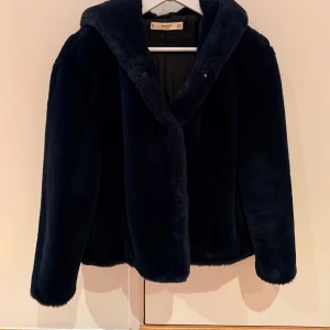 Mörkblå fuskpälsjacka med huva från Mango - Säljer en mörkblå fuskpälsjacka från Mango med stor, mysig huva. Jackan har en loose passform och är tillverkad i mjukt syntetmaterial som ger en lyxig känsla. Perfekt för kalla dagar och har en stilren look utan synliga detaljer på framsidan.