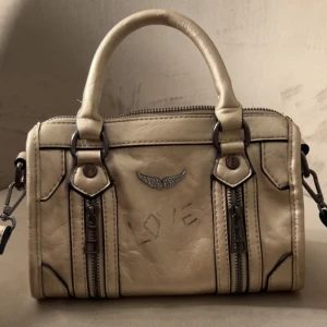 Guld Zadig liknande väska - Cool guld handväska i skinnimitation med två rejäla handtag och avtagbar axelrem. Väskan har dekorativa dragkedjor framtill, metallfärgade detaljer och ett emblem med vingar. Snygg boxig form och texten 'LOVE' på framsidan.