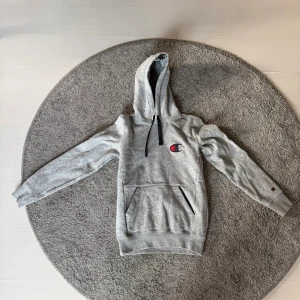 Grå hoodie från Champion - Grå hoodie från Champion med svart snörning och stor logga på bröstet. Klassisk känguruficka framtill och ribbade muddar vid ärmslut och nederkant. Perfekt för en avslappnad stil.