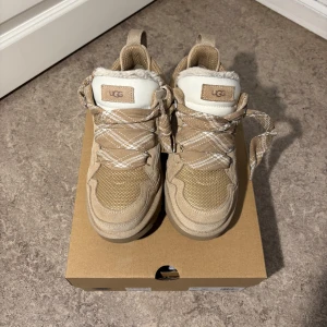 Lowmels Uggs sand  - Säljer ett par snygga beige sneakers från UGG i modell Lowmel Storlek 39 (EU). Skorna har rutiga snören, meshdetaljer och är fodrade med mjuk fuskpäls för extra komfort. Kommer med ett extra par skorsnören, har även kvar kartongen. Köpta för en månad sen, är i väldigt bra skick! Kan skicka fler bilder ifall det önskas. Buda på! 