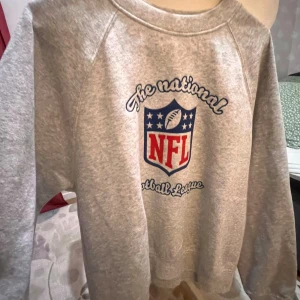 Grå NFL sweatshirt från Divided S - Säljer en grå sweatshirt från Divided i storlek S med stort NFL-tryck i rött och blått framtill. Tröjan har rund hals, långa ärmar och mjukt material som känns skönt mot huden. Perfekt för dig som gillar sportig stil och vill sticka ut med en cool logga.