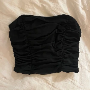 Svart tubtopp från Forever 21 - Svart tubtopp från Forever 21 med snygga rynkade detaljer framtill. Toppen är axelbandslös och har en tight passform som sitter på plats. Köpt i New York för två år sedan så kommer inte ihåg priset. Men använd ensast ett fåtal gånger💋 