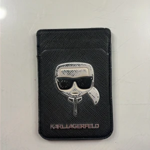 Svart plånbok från Karl Lagerfeld - Slimmad svart plånbok från Karl Lagerfeld med ikonisk silverfärgad detalj på framsidan och diskret logga. Plånboken har plats för kort och ett smart fönster på baksidan för enkel åtkomst. Snygg och stilren design som passar perfekt i fickan. Den är även magnetisk.