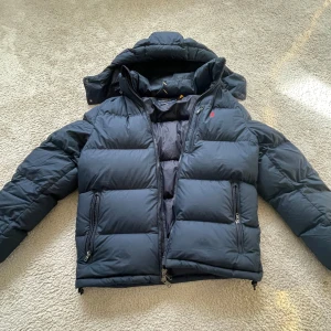 Svart dunjacka från Polo Ralph Lauren - Snygg marinblå dunjacka från Polo Ralph Lauren med röd broderad logga på bröstet. Jackan har huva, två dragkedjefickor och kraftig vaddering som håller dig varm. Klassisk pufferstil med raka linjer och dragkedja framtill.