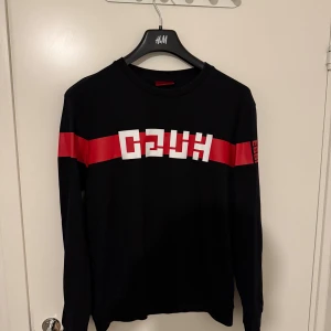 Svart långärmad tröja med röd rand (Hugo) - Svart långärmad tröja från hugo med en bred röd rand över bröstet och ärmarna. Framsidan har ett grafiskt vitt tryck och en liten röd detalj på ärmen. Tröjan har rund hals och är i mjuk bomull, perfekt för en cool streetstyle.