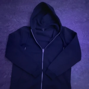 Svart hoodie med dragkedja från Smog - Svart hoodie från Smog i storlek S med regular fit. Tröjan har huva med snörning, långa ärmar och en praktisk dragkedja framtill. Tillverkad i mjukt bomullsmaterial, perfekt för chill dagar. Snygg och enkel design som funkar till det mesta.