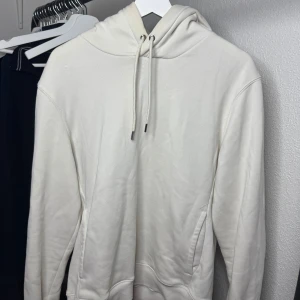 Vit hoodie från H&M i storlek S - Säljer en stilren vit hoodie från H&M i storlek S. Tröjan har huva med snörning och en klassisk känguruficka framtill. Perfekt för dig som gillar en enkel och clean look. Materialet är mjukt och bekvämt, troligtvis bomullsmix.