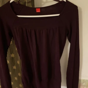 Mörklila långärmad tröja från Esprit - Mörklila långärmad tröja från Esprit med bred, ribbad kant vid halsringningen och nedtill. Tröjan har en lätt rynkad detalj framtill och är tillverkad i mjukt material som känns skönt mot huden. Perfekt för lager-på-lager under kyligare dagar.