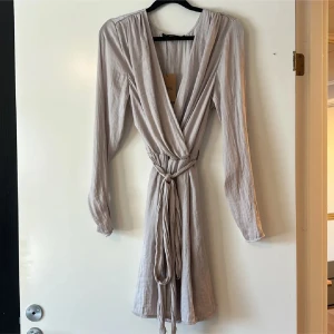 Sommarklänning  - Supersnygg beige kort omlottklänning med långa ärmar och knytband i midjan. Klänningen har v-ringning och är tillverkad i ett lätt, skrynkligt material som ger en avslappnad men chic look. 