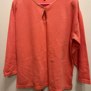 Korallfärgad långärmad tröja - Säljer en korallfärgad långärmad tröja med rund hals och snygg knytning vid halsringningen. Tröjan är tillverkad i ett mjukt bomullsmaterial och har en loose passform som gör den skön att bära. Perfekt för dig som gillar enkel och stilren design.