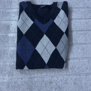 Mörkblå argyle tröja från Skill - Snygg mörkblå tröja från Skill med klassiskt argylemönster i blått, grått och beige. V-ringad hals och långa ärmar, perfekt för dig som gillar preppy stil. Materialet är mjukt och känns skönt mot huden. Passar dig som vill sticka ut med stilrena detaljer.